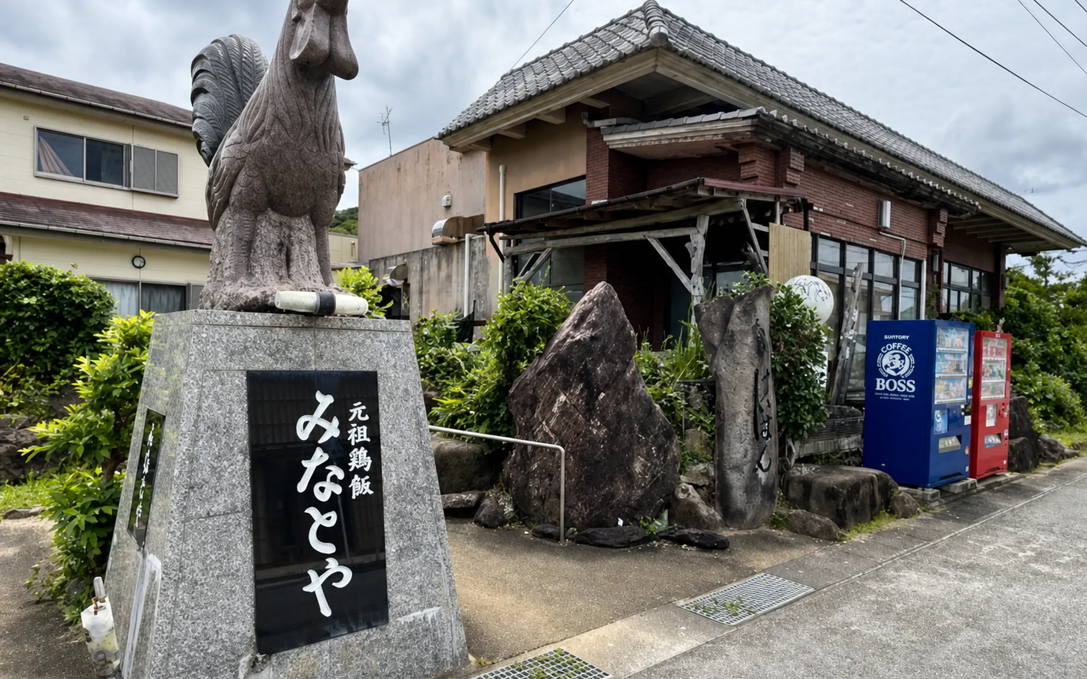 鶏の石像と「元祖鶏飯 みなとや」の石碑が立つ店先。奥に瓦屋根の店舗と自動販売機