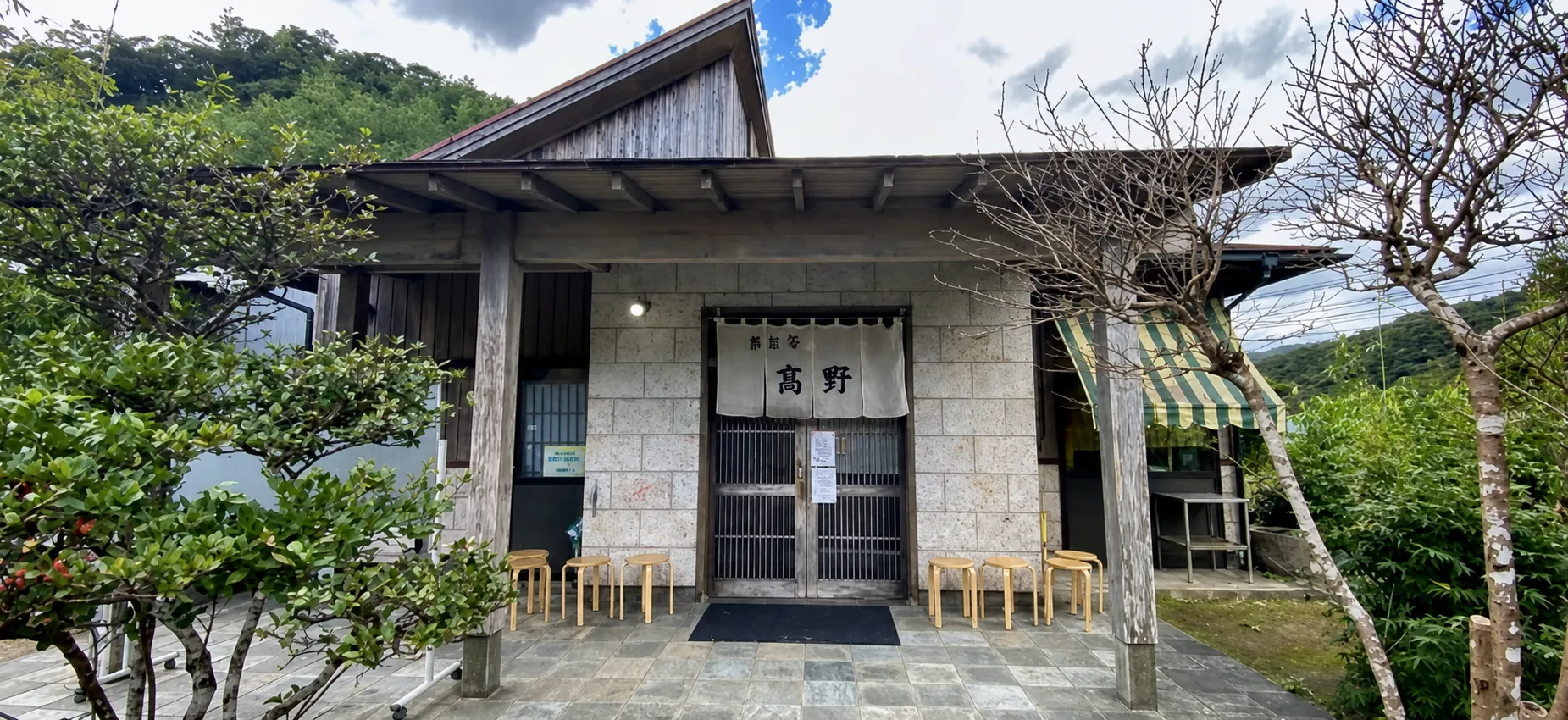 石積みの壁に「髙野」の白い暖簾がかかる島とうふ屋の入口。引き戸の前に木のスツールが並ぶ