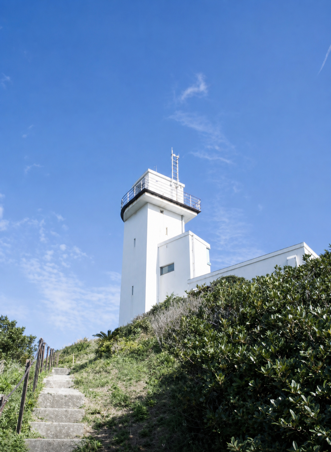 石段の先に立つ笠利崎灯台。青空と緑の茂みに囲まれた白い灯台
