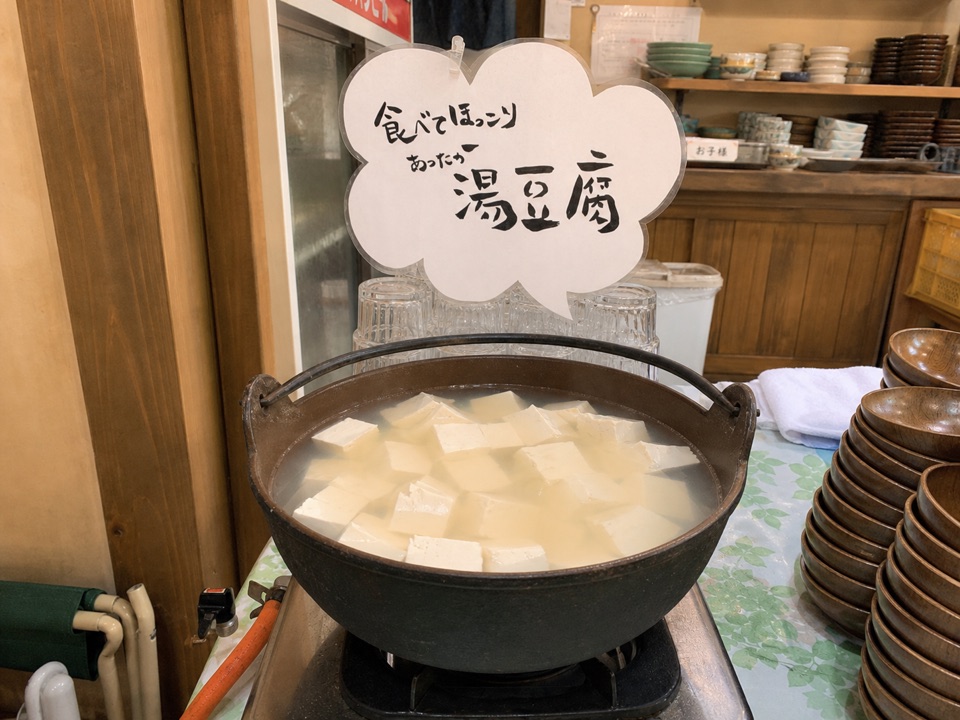 「食べてほっこり あったか湯豆腐」と手書きされた雲形のPOPを背に、カセットコンロにかけられた大きな土鍋。角切りの豆腐が白濁した湯の中いっぱいに煮えている